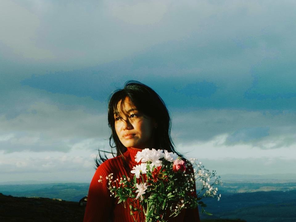 Still from Jolie: a woman in a red jumper holds white flowers against the hills.
